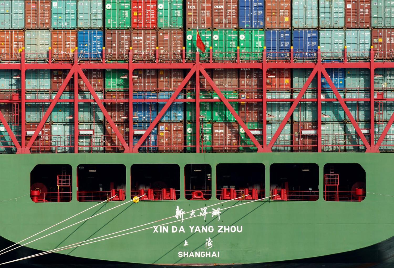 Shipping containers stacked on a ship's deck, behind a red barricade flying the flag of China